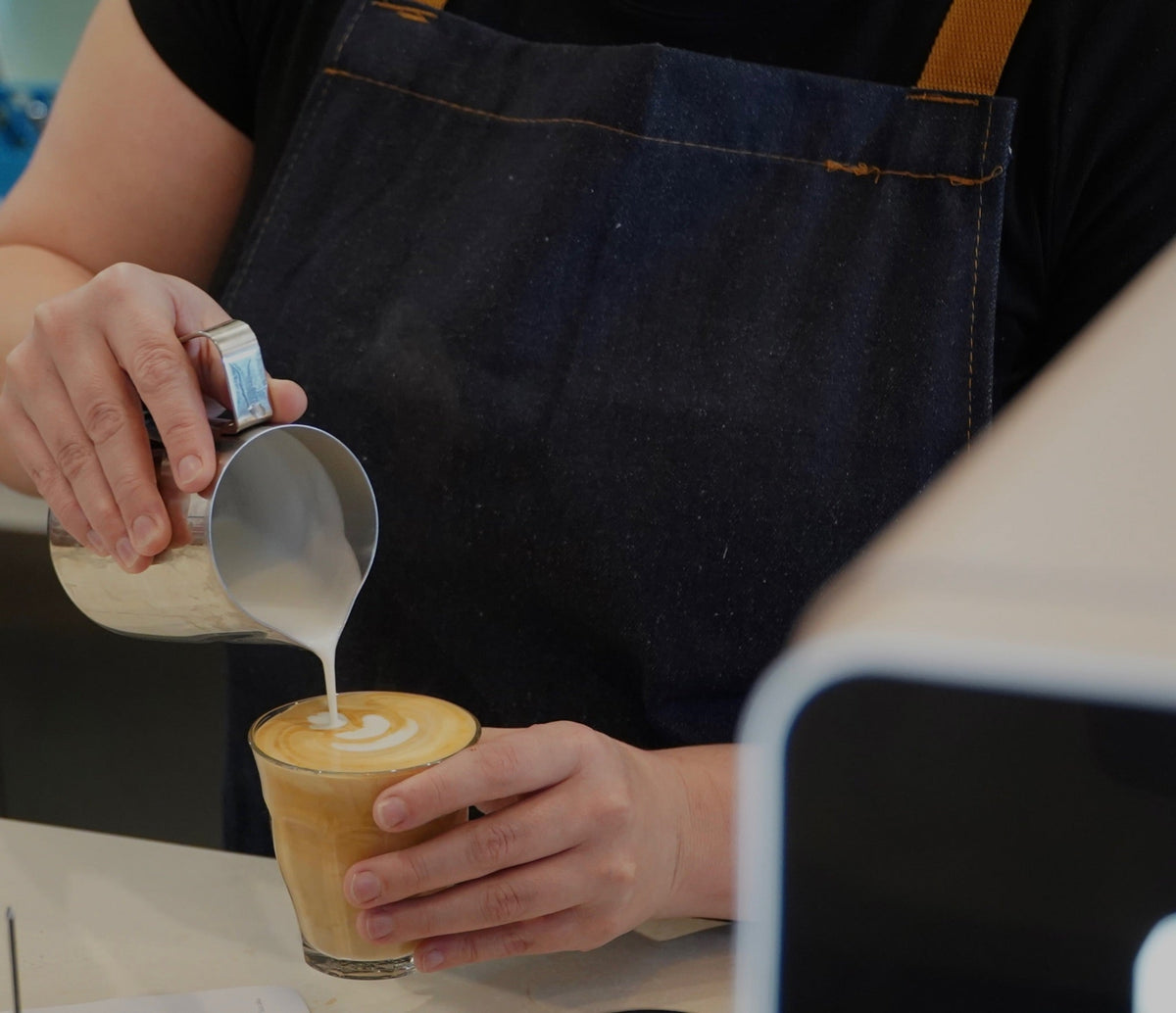 A close-up of a barista pouring steamed milk from a metal pitcher into a glass cup of latte art, with the person wearing a dark denim apron.